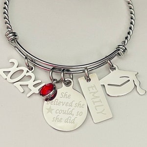 May include: Silver-toned graduation bracelet with charms. Charms include the year 2024, a graduation cap, a tag with "EMILY" engraved, and a circular charm with the text "She believed she could, so she did." A red bead is also included.