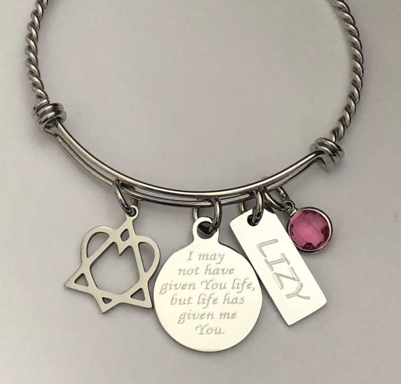 May include: A silver bangle bracelet with a heart charm, a round charm with the inscription "I may not have given You life, but life has given me You.", a tag charm with the name "LIZY" engraved on it, and a pink crystal charm.