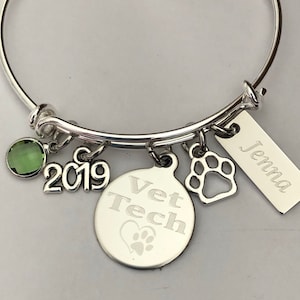 May include: Silver charm bracelet with a green gemstone, a charm that says "Vet Tech" with a paw print, a charm that says "2019", a paw print charm, and a tag that says "Jenna".