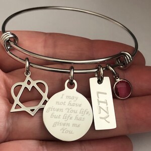 May include: A silver expandable bangle bracelet with four charms. The charms include a heart with a star inside, a round charm with the engraved text "I may not have given you life, but life has given me you", a rectangular charm with the name "LIZY" engraved on it, and a small red crystal charm.
