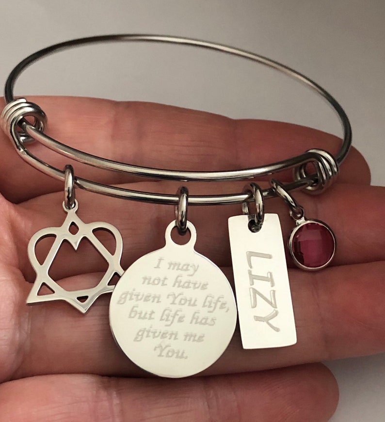 May include: A silver expandable bangle bracelet with four charms. The charms include a heart with a star inside, a round charm with the engraved text "I may not have given you life, but life has given me you", a rectangular charm with the name "LIZY" engraved on it, and a small red crystal charm.