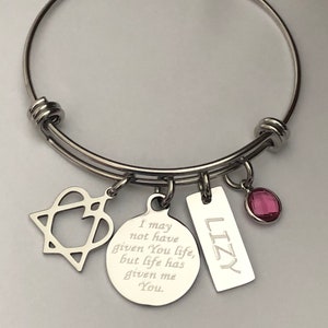 May include: A silver bangle bracelet with charms. The charms include a heart with a star inside, a round charm with the text "I may not have given You life, but life has given me You.", a rectangular charm with the text "LIZY", and a pink crystal charm.