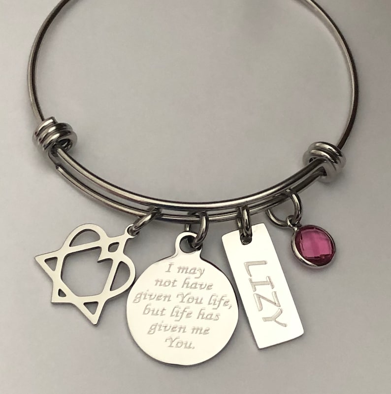 May include: A silver bangle bracelet with charms. The charms include a heart with a star inside, a round charm with the text "I may not have given You life, but life has given me You.", a rectangular charm with the text "LIZY", and a pink crystal charm.