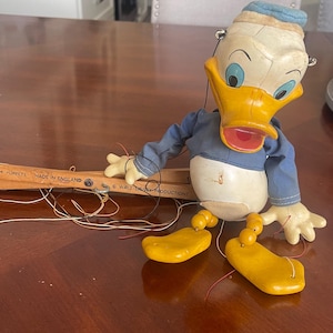 May include: A vintage Donald Duck marionette puppet featuring a white and yellow molded head with a blue cap, a blue fabric shirt, and yellow wooden feet. The puppet is attached to a wooden control bar marked with "MADE IN ENGLAND" and "WALT DISNEY PRODUCTIONS" text, connected by thin black and red strings.