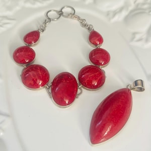 May include: A silver bracelet featuring multiple round and teardrop-shaped red stones in polished settings, accompanied by a matching elongated marquise-shaped red stone pendant with a silver bail.