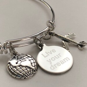 May include: Silver charm bracelet with three charms: a globe, an airplane, and a round charm with the words "Live your dream" engraved on it.