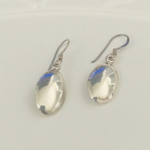 May include: A pair of silver drop earrings. Each earring features a polished, oval-shaped design with a hook closure. The earrings reflect light, creating a bright, shiny appearance. The earrings are displayed against a white background.