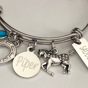 May include: Silver charm bracelet with a blue gem, horseshoe, horse charm, and a tag that says "Saddle Up" and a round tag with the name "Piper" engraved on it.