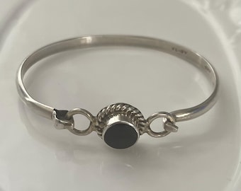 May include: A silver bangle bracelet with a black onyx stone set in a silver bezel. The bracelet has a simple design with a single stone and two small silver loops.