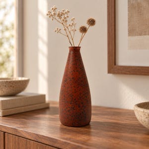 May include: A tall, tapered ceramic vase with a matte, burnt-orange finish and dark brown speckled texture. The vase has a narrow neck and a wide, rounded base. It contains a small arrangement of dried, light-colored botanical stems.