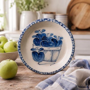 May include: A round ceramic bowl with an off-white base and a blue hand-painted design. The center features a basket filled with blue apples, while the rim is decorated with a repeating blue checkered pattern. The bowl has a textured surface.