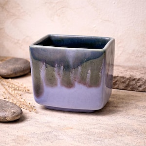 May include: A square ceramic planter with a gradient glaze in shades of blue, green, and lavender. The planter has a slightly rounded base and a dark blue interior. The glaze creates a textured, dripped effect. Ideal for small plants or succulents.