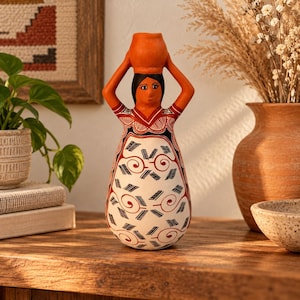 May include: A terracotta-colored ceramic figurine of a person carrying a vessel on their head. The figure features a white base with intricate black and red geometric patterns, including swirls and hatch marks, across the body and arms.