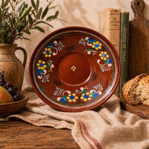 May include: A round, brown ceramic bowl featuring a hand-painted floral pattern in yellow, blue, and white. The centre of the bowl displays a small cluster of five yellow dots, while the rim is decorated with repeating floral motifs and cross-hatch marks.