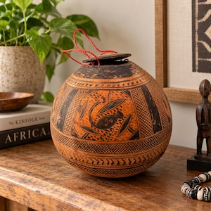Puede incluir: Un recipiente esférico de calabaza en tonos naranja con complejos patrones grabados en negro, incluyendo sombreado geométrico y motivos de aves estilizadas. El contenedor tiene una tapa plana oscura y un lazo de hilo rojo en la parte superior.