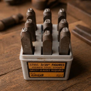 Puede incluir: Un juego de nueve sellos de acero metálico dispuestos en un soporte de plástico gris. La etiqueta frontal dice 22901 3/16" FIGURES PREMIER STEEL STAMPS SAFETY GLASSES REQUIRED, The C.H. Hanson Company, MFG. 690211. Cada sello tiene un carácter arriba.