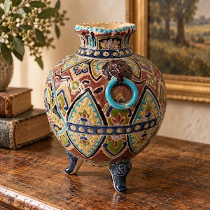 May include: A round ceramic vase with three dark blue feet and a scalloped rim. The surface features a brown base with intricate geometric patterns in green, yellow, and blue. A dark brown lion head handle holds a bright blue ring.