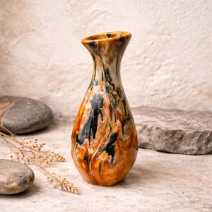 Może przedstawiać: Ceramiczny wazon o unikalnym, organicznym designie. Wazon charakteryzuje się mieszanką ciepłych kolorów, w tym pomarańczowego, brązowego i czarnego, z błyszczącym wykończeniem. Kształt jest wydłużony z rozszerzonym wierzchołkiem, odpowiedni do eksponowania kwiatów lub jako element dekoracyjny.