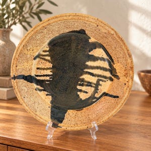 May include: A round ceramic plate with a speckled tan and beige base featuring a prominent, dark, abstract brushstroke design in the center. The plate has a raised rim and is displayed on a clear plastic stand, showcasing its textured surface.