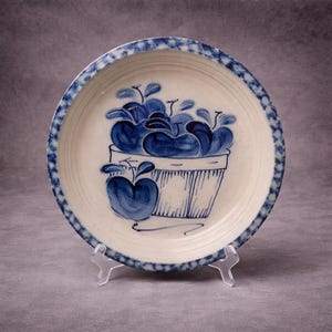 Puede incluir: Un plato de cerámica con un centro color crema y un borde azul con un patrón de puntos. El centro del plato muestra una ilustración azul de manzanas en una cesta. El plato se exhibe sobre un soporte transparente.