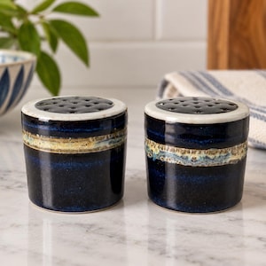 May include: Two cylindrical ceramic shakers featuring a deep navy blue glaze with a horizontal band of textured, mottled beige and light blue detailing around the center. Each shaker has a flat, white-rimmed top with multiple small holes.