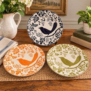 May include: Three round ceramic plates featuring a bird perched on a branch surrounded by floral and pinecone patterns. One plate is dark blue on white, one is orange on white, and one is olive green on white, all with a consistent design.
