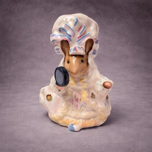 May include: A ceramic figurine of a mouse wearing a white bonnet with blue and pink accents, and a matching robe. The mouse is holding a small, round, blue mirror. The figurine has a painted floral design.