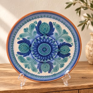 May include: A round ceramic plate with a terracotta rim and a cream-colored center. The surface features a symmetrical, intricate floral pattern in shades of deep navy blue, bright blue, and teal, surrounded by a dotted blue border.