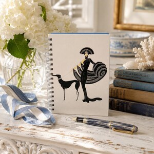 May include: A spiral-bound notebook featuring a graphic illustration of a person in a long black gown and a large headpiece walking a black greyhound on a leash. The cover is off-white with a clean, centered design and a silver metal spiral binding.