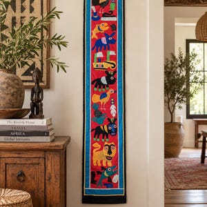 May include: A long, vertical textile wall hanging featuring a bright red center panel with colorful embroidered animal and boat motifs, framed by a blue border and a thin black edge. The design includes stylized figures of birds, fish, and mammals.