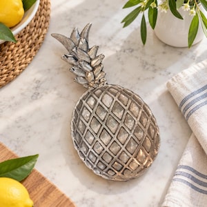May include: A silver-toned metal tray shaped like a pineapple, featuring a textured diamond-patterned body and a detailed leafy top. The tray has a metallic finish with subtle dark shading in the crevices, creating a realistic pineapple appearance.