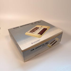 Puede incluir: Una caja de cartón rectangular para una unidad Commodore C2N Datasette modelo 1530. La caja presenta un diseño en azul claro y blanco con líneas horizontales, el logotipo de Commodore Computer y una imagen impresa de la unidad de casete beige.
