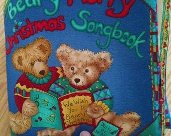 Pode incluir: Um livro de tecido azul com o título "Beary Merry Christmas Songbook". Dois ursos de peluche marrons cantam de um livro com notas musicais. O texto "We Wish You A Beary Christmas" está no livro.