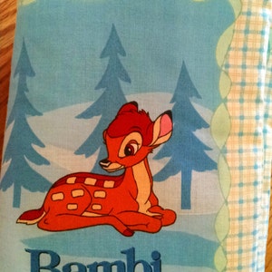May include: A blue and white checkered baby blanket with a cartoon image of Bambi the deer in a forest setting. The blanket has the word "Bambi" embroidered in blue on the bottom.