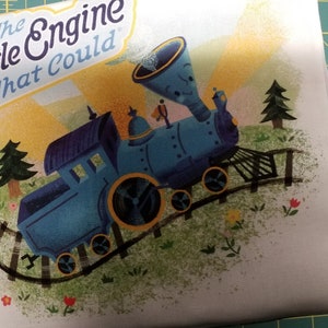 Könnte beinhalten: Eine blaue Lokomotive mit einer gelben Pfeife und den Worten "The Little Engine That Could" an der Seite. Der Zug befindet sich auf einem Gleis mit grünem Gras und Blumen.