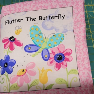Könnte beinhalten: Ein rosa Stoffbuch mit dem Titel "Flutter The Butterfly" auf dem vorderen Umschlag. Das Buch zeigt einen bunten Schmetterling mit blauen Flügeln, gelbem und grünem Körper und schwarzen Punkten. Der Schmetterling fliegt über einem Feld mit rosa, blauen und gelben Blumen.