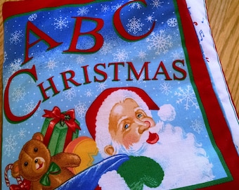 Pode incluir: Um livro de tecido azul e vermelho com o texto "ABC Christmas" e uma imagem do Papai Noel, um urso de pelúcia e presentes.