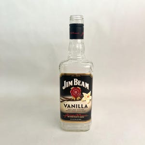 Puede incluir: Una botella de vidrio transparente de Jim Beam Vanilla con una etiqueta negra y dorada. La etiqueta presenta el logotipo de Jim Beam, la palabra "VANILLA" y una ilustración de flor de vainilla. La botella está vacía.