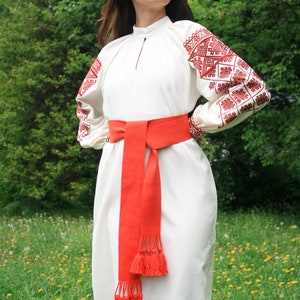 Pode incluir: Uma mulher vestindo um vestido branco bordado com detalhes vermelhos e uma faixa vermelha com borlas. Ela está em pé em um campo de grama verde e flores amarelas.