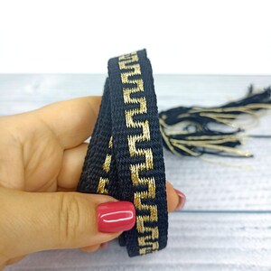 May include: Black and gold trim with a geometric pattern. The trim is woven and has a tassel on the end.