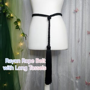 Könnte beinhalten: Ein schwarzer Rayon-Seilgürtel mit langen Quasten wird auf einer Schaufensterpuppe gezeigt. Der Gürtel ist in der Taille gebunden und hat eine lange, dekorative Quaste. Der Text "Rayon Rope Belt with Long Tassels" ist im Bild zu sehen.