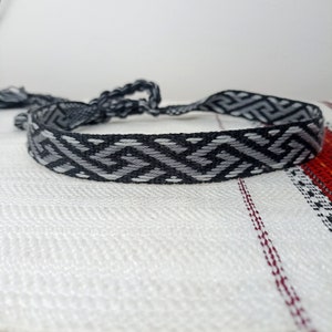 May include: A woven bracelet with a geometric pattern in shades of gray and black. The bracelet is displayed on a white and red patterned surface. The ends of the bracelet are tied together.