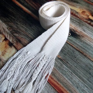May include: White woven sash with long fringe tassels. The sash is folded and tied in a knot. The background is a wooden surface.