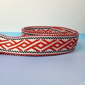 K&ouml;nnte beinhalten: Rotes, wei&szlig;es und schwarzes gewebtes Band mit geometrischem Muster. Das Band ist aufgerollt und hat eine strukturierte Oberfl&auml;che.
