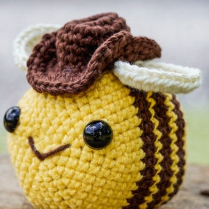 May include: A crocheted bee wearing a brown cowboy hat with white accents. The bee has a yellow body with brown stripes and black eyes.