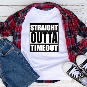May include: White t-shirt with a black graphic that says "STRAIGHT OUTTA TIMEOUT". The shirt is laid out on a white wooden surface with a red and black plaid shirt, blue jeans, and black sneakers.