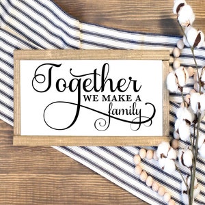 May include: A wooden sign with the text "Together we make a family" in black cursive font. The sign is on a wooden surface with a white and blue striped fabric and a sprig of cotton.