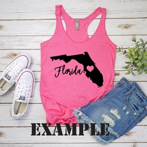 May include: Pink tank top featuring a black outline of the state of Florida with the word "Florida" in black script. A small pink heart is inside the outline of the state. The tank top is lying on a white wooden surface with a pair of white trainers and a pair of blue denim shorts.
