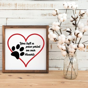 May include: A wooden framed sign with a white background and a red heart with a black paw print inside. The text on the sign reads "You left a paw print on our hearts".
