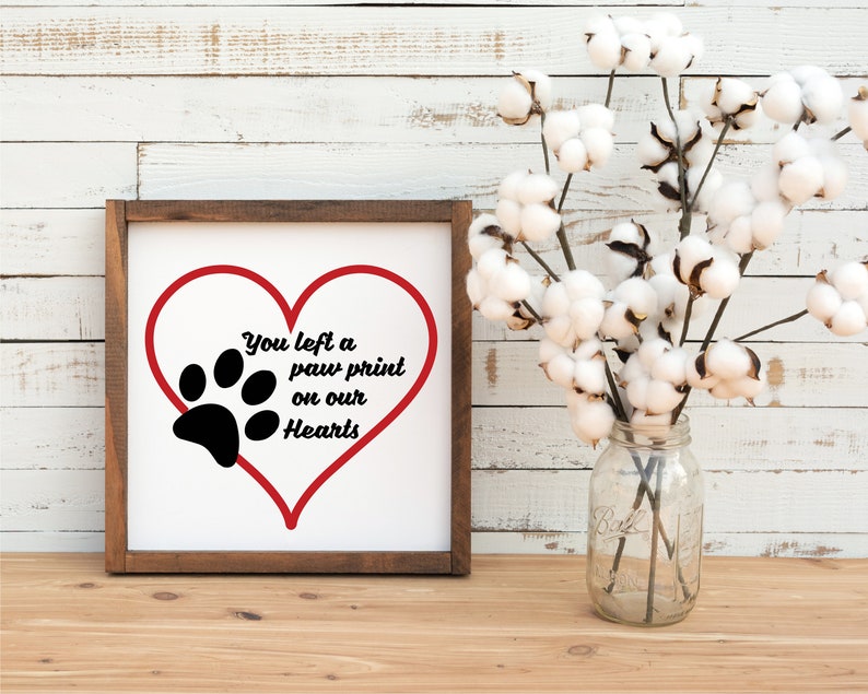 May include: A wooden framed sign with a white background and a red heart with a black paw print inside. The text on the sign reads "You left a paw print on our hearts".